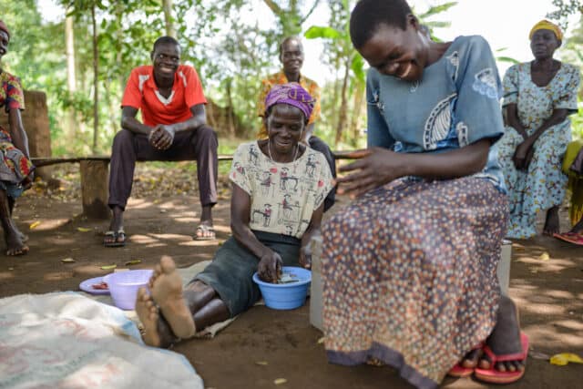 A group of people sat outside at a Village Savings and Loan Associations meeting; a woman holds a bowl filled with cash.