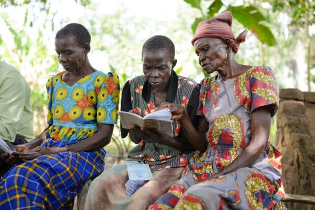 Three women sitting outdoors looking at notebooks at a Village Savings and Loan Associations meeting.